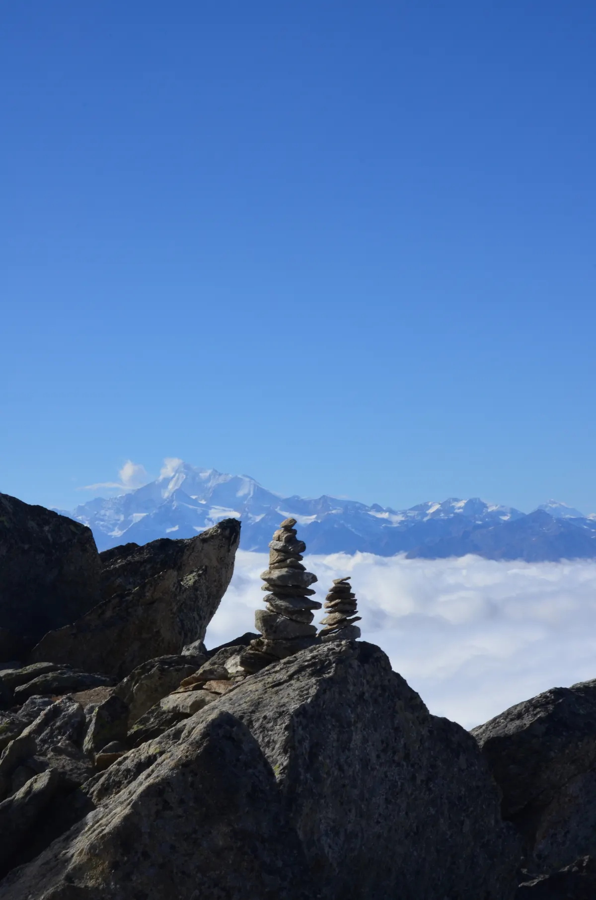 Deux cairns en équilibre sur un rocher, au-dessus d’une mer de brouillard face aux sommets valaisans.